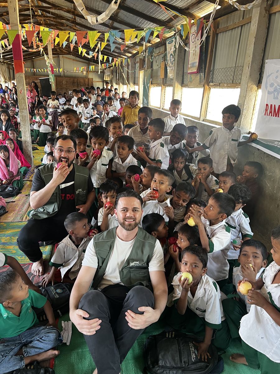 Zwei Vuslat-Helfer inmitten vieler Schulkinder, die Äpfel essen, im Rohingya-Camp